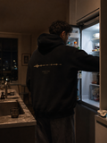 Person wearing the black EVAREN Room Tone Hoodie from behind in a late-night kitchen, showing the muted-gold waveform back artwork.