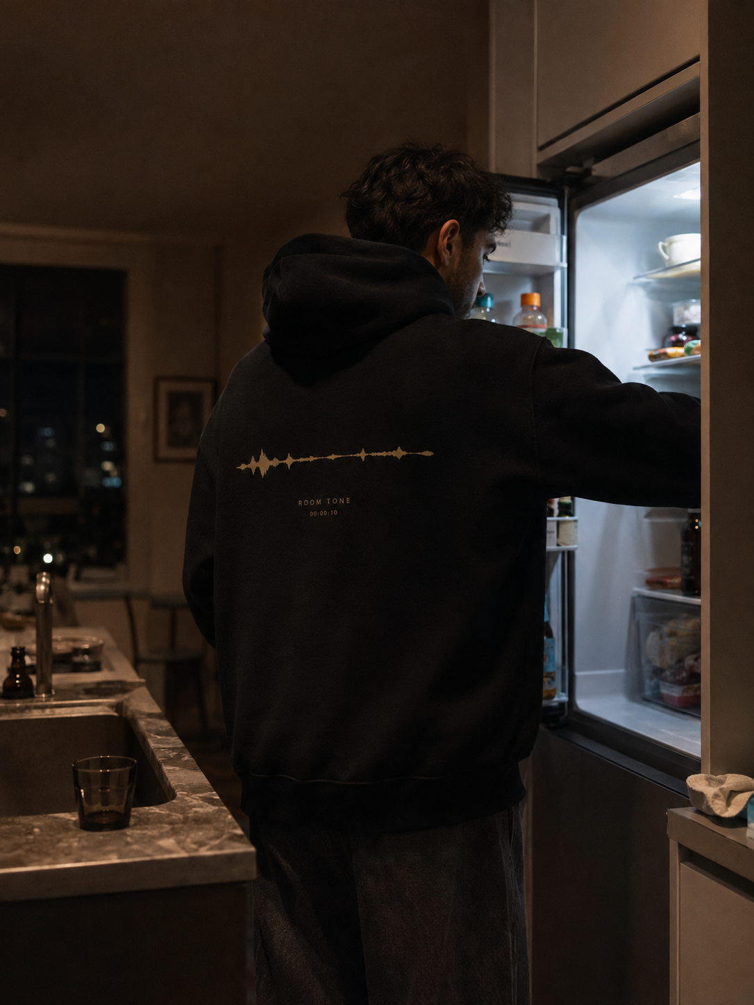 Person wearing the black EVAREN Room Tone Hoodie from behind in a late-night kitchen, showing the muted-gold waveform back artwork.