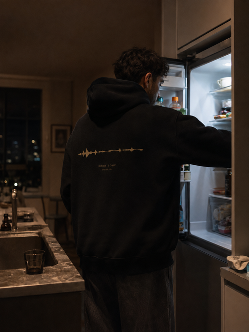 Person wearing the black EVAREN Room Tone Hoodie from behind in a late-night kitchen, showing the muted-gold waveform back artwork.