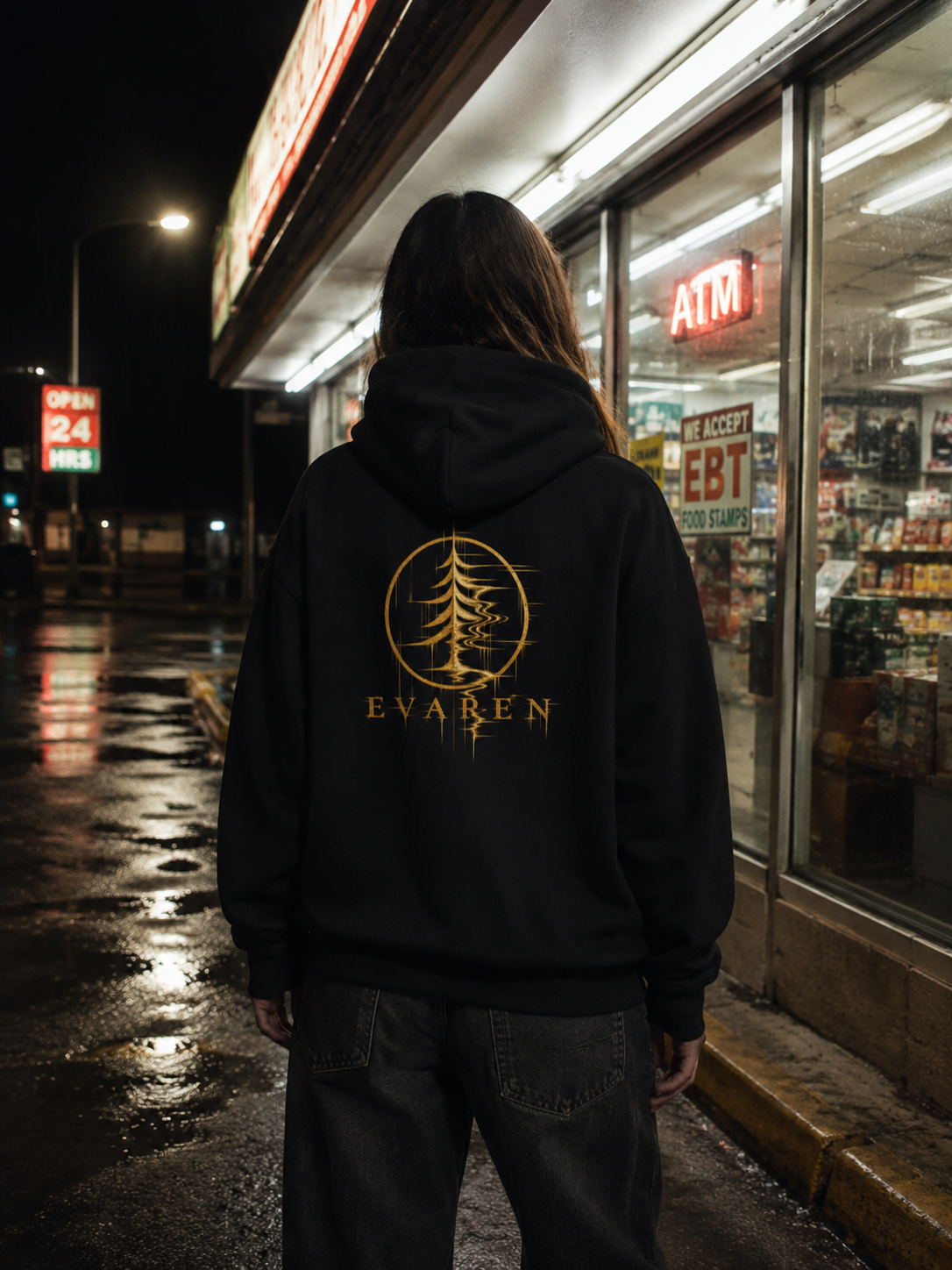 Person wearing The Distortion Hoodie by EVAREN outside a convenience store at night, showing the gold distorted evergreen back graphic.