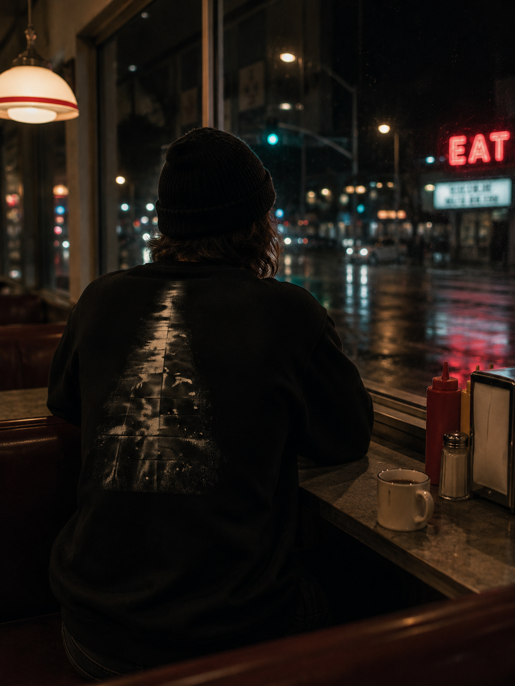 Person wearing The Witness Sweatshirt at a late-night diner window, with the abstract evergreen back graphic visible against a rainy city street.