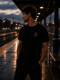     Man wearing black EVAREN Signature Tee with gold tree emblem on a wet train platform at dusk