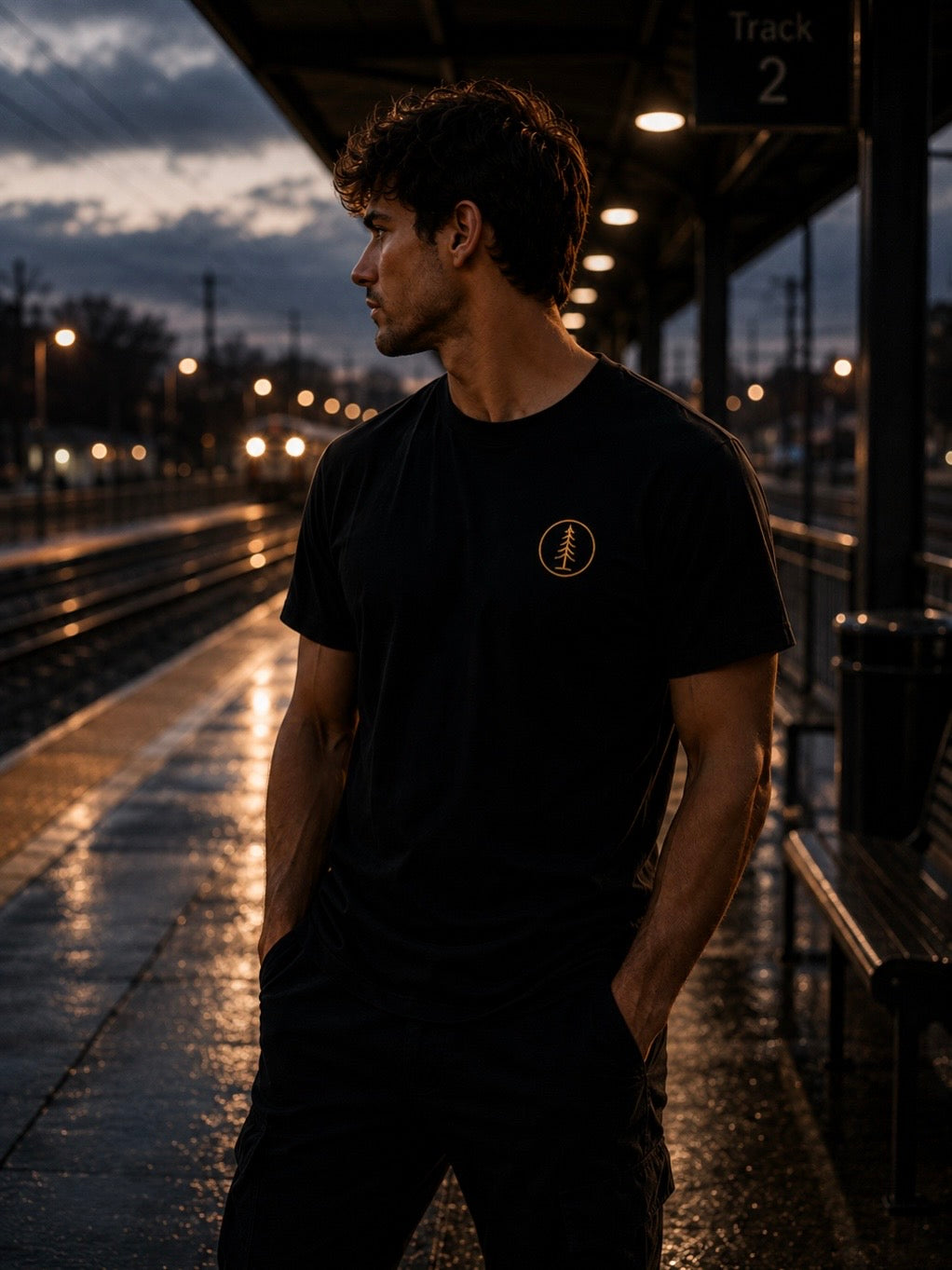     Man wearing black EVAREN Signature Tee with gold tree emblem on a wet train platform at dusk