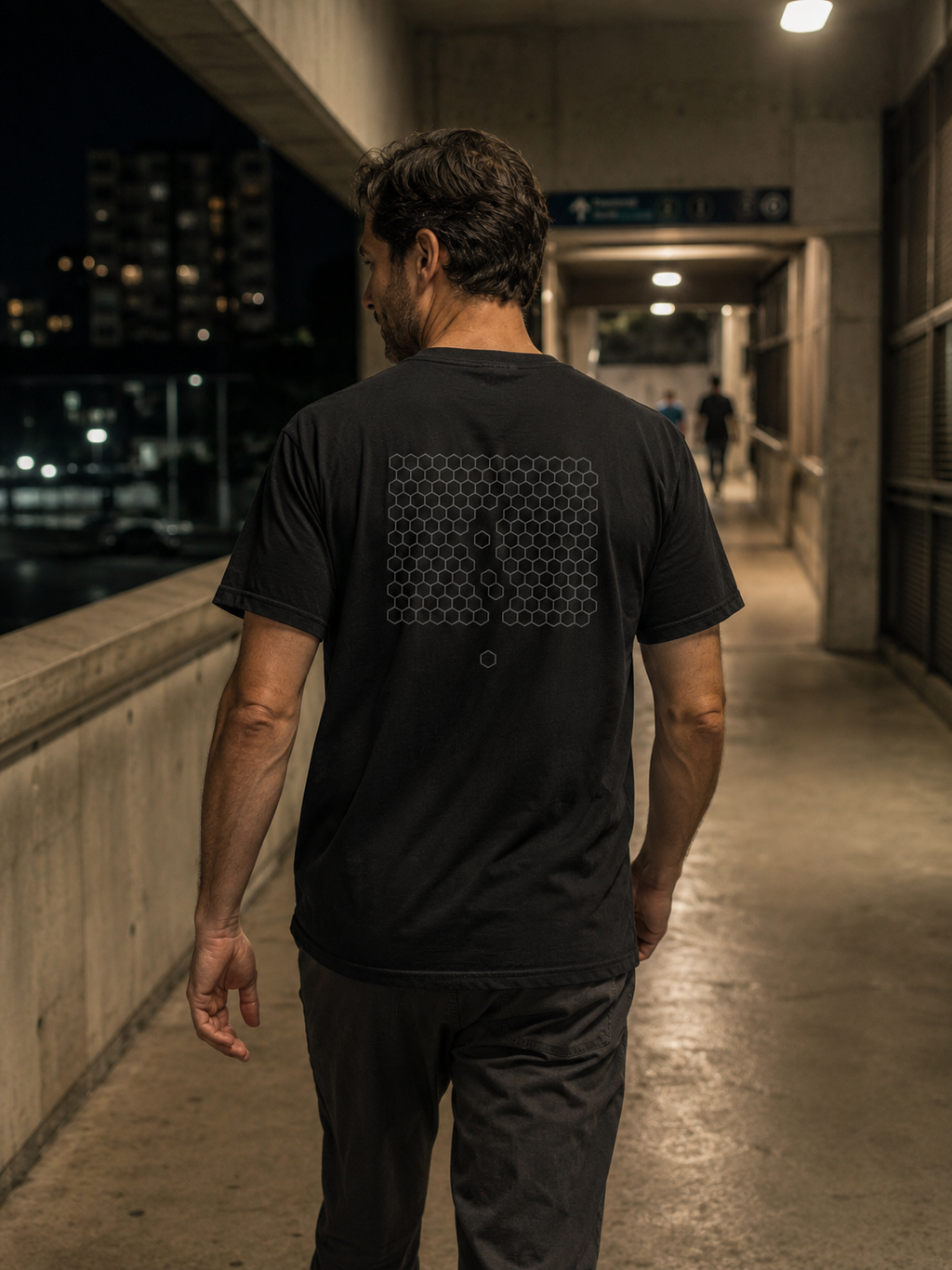 Man wearing The Way Through Tee from behind on a nighttime urban walkway, showing the subtle geometric back graphic.