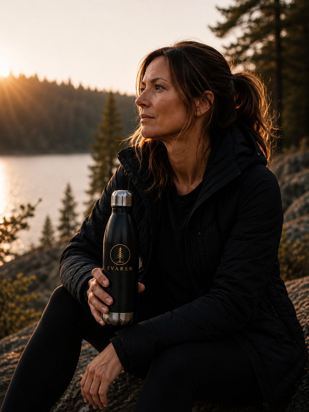 Woman holding a black EVAREN stainless steel water bottle beside a mountain lake at sunset