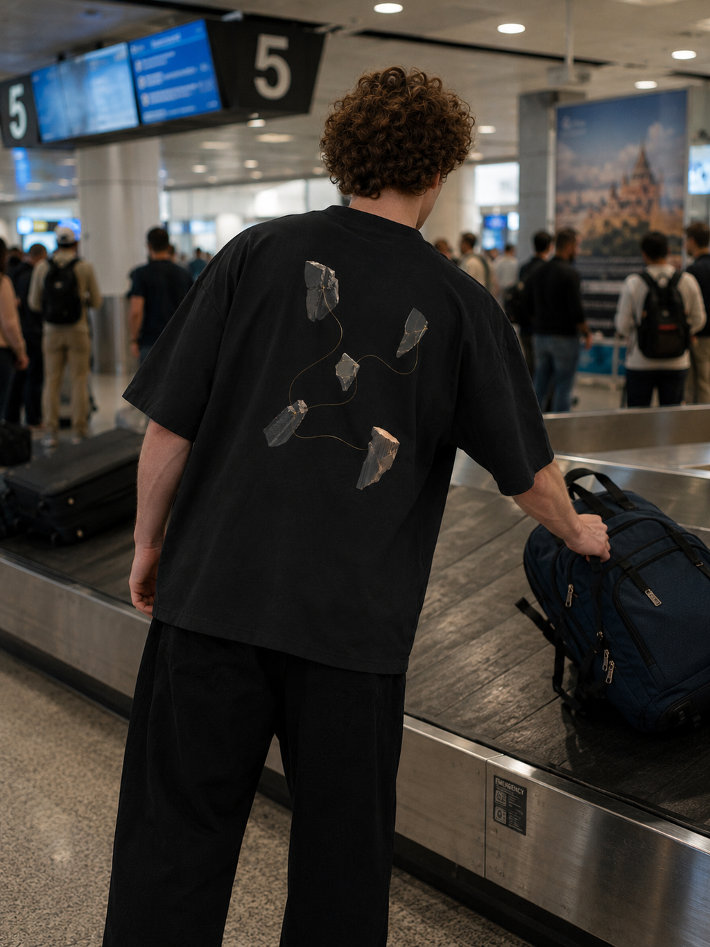 The Thread Remains Tee worn at an airport baggage claim, showing the large back graphic of fractured forms connected by a fine gold thread.