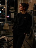 Woman wearing a black EVAREN Signature Sweatshirt with gold tree emblem in a nighttime laundromat.