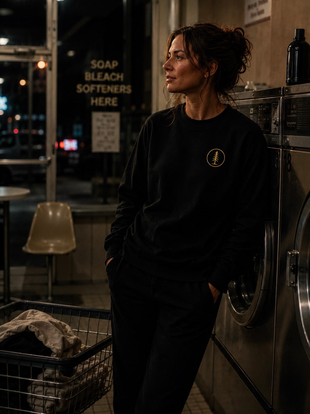 Woman wearing a black EVAREN Signature Sweatshirt with gold tree emblem in a nighttime laundromat.