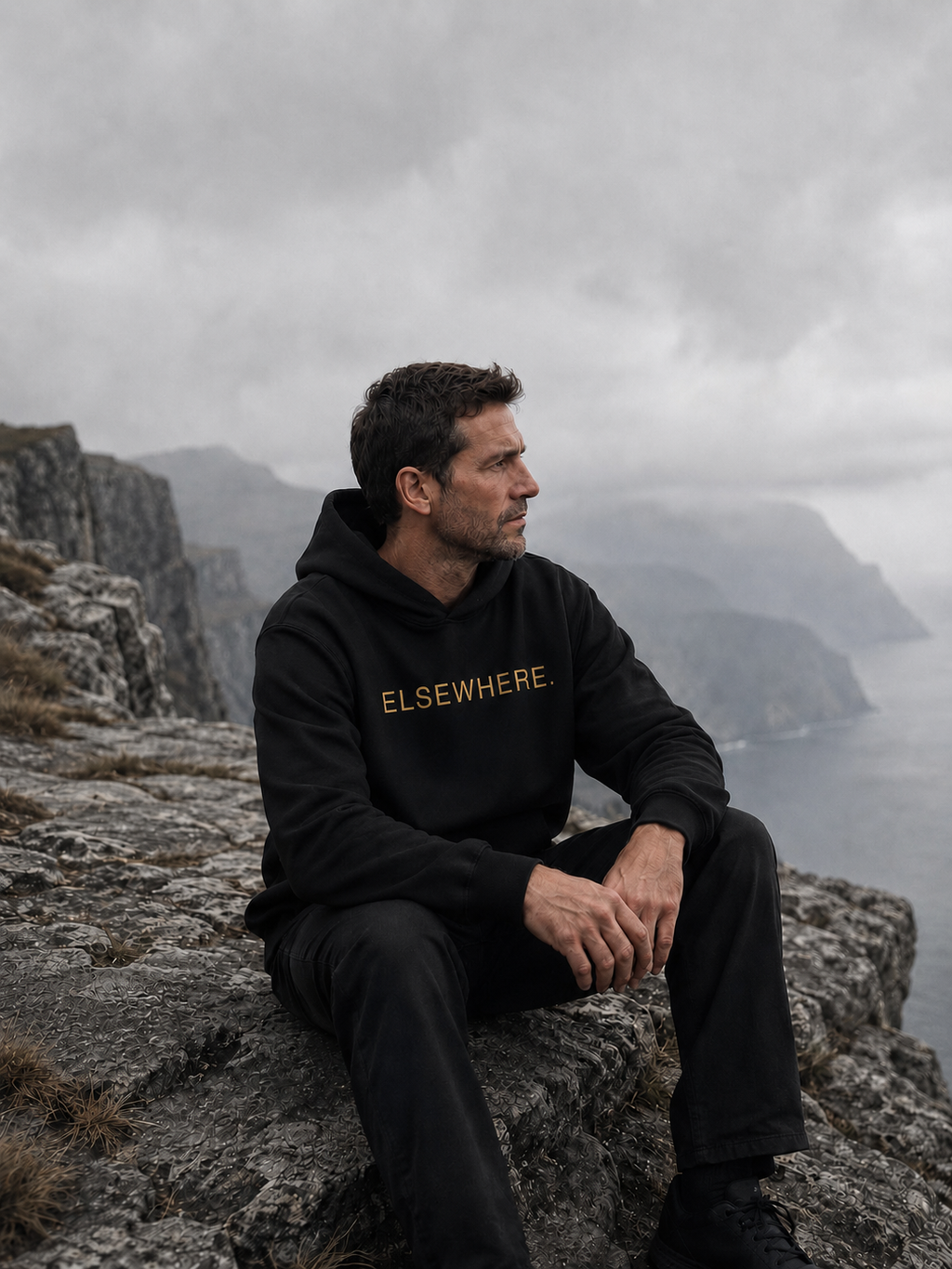 Man wearing The Elsewhere Hoodie seated on a rocky overlook above misty coastal cliffs.