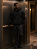 Man wearing the black EVAREN Signature Hoodie with a gold embroidered evergreen emblem in a warmly lit elevator.