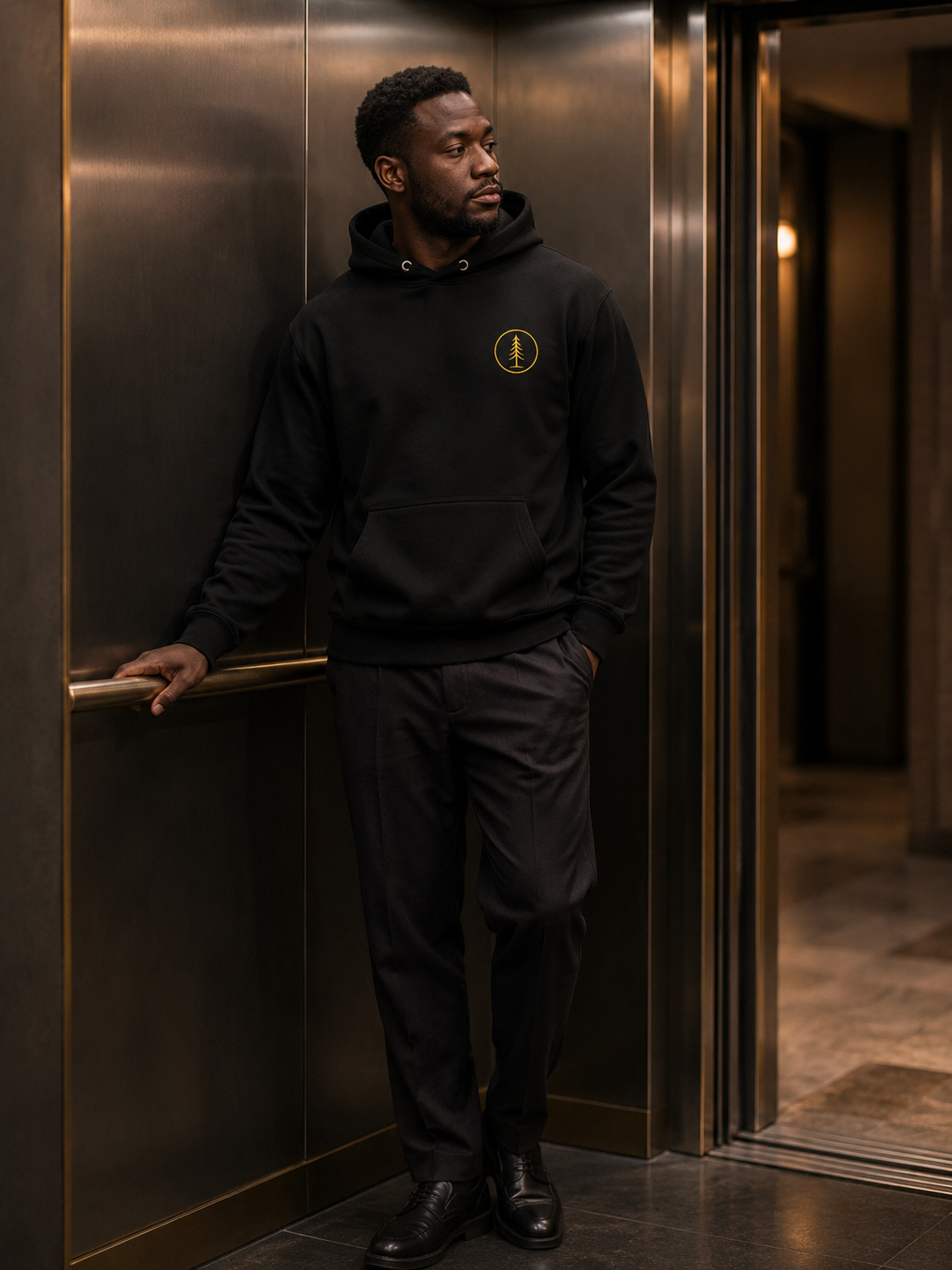 Man wearing the black EVAREN Signature Hoodie with a gold embroidered evergreen emblem in a warmly lit elevator.