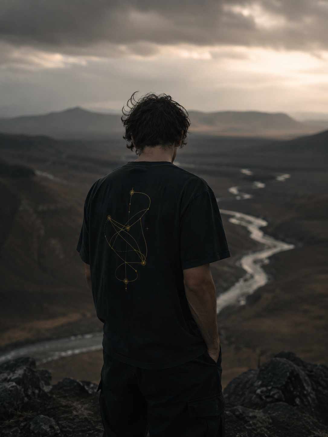 Person wearing The Nonlinear Tee overlooking a winding river valley at dusk, showing the gold nonlinear path graphic on the back.