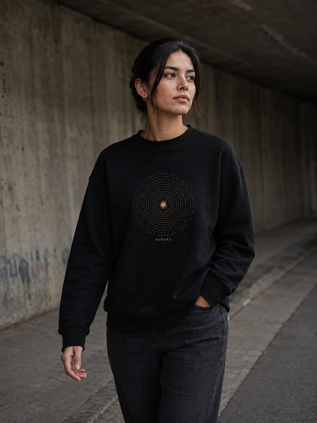 Woman wearing The Echoes Sweatshirt in black while walking through a concrete underpass, showing the antique-gold concentric ring front design.