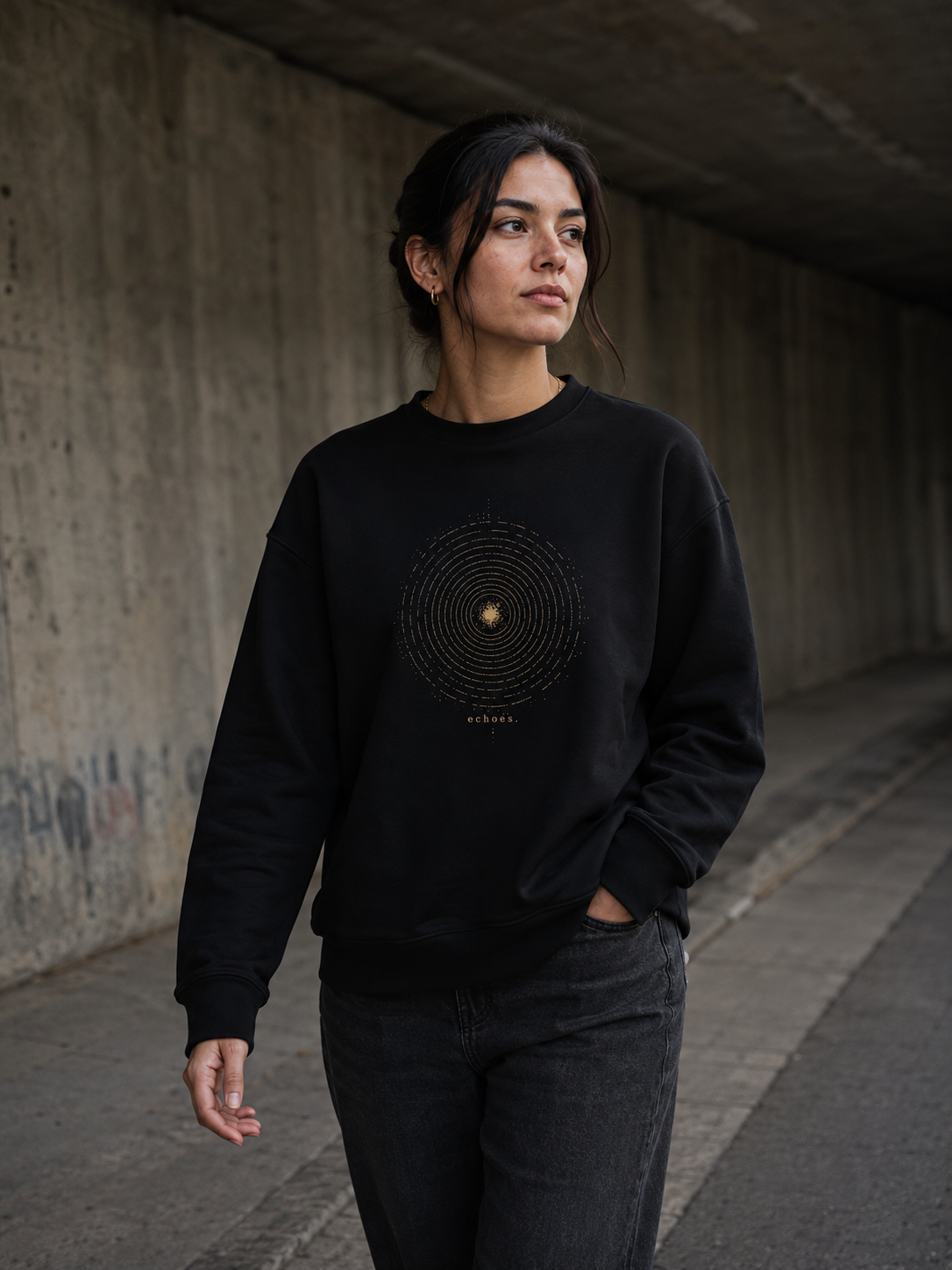 Woman wearing The Echoes Sweatshirt in black while walking through a concrete underpass, showing the antique-gold concentric ring front design.