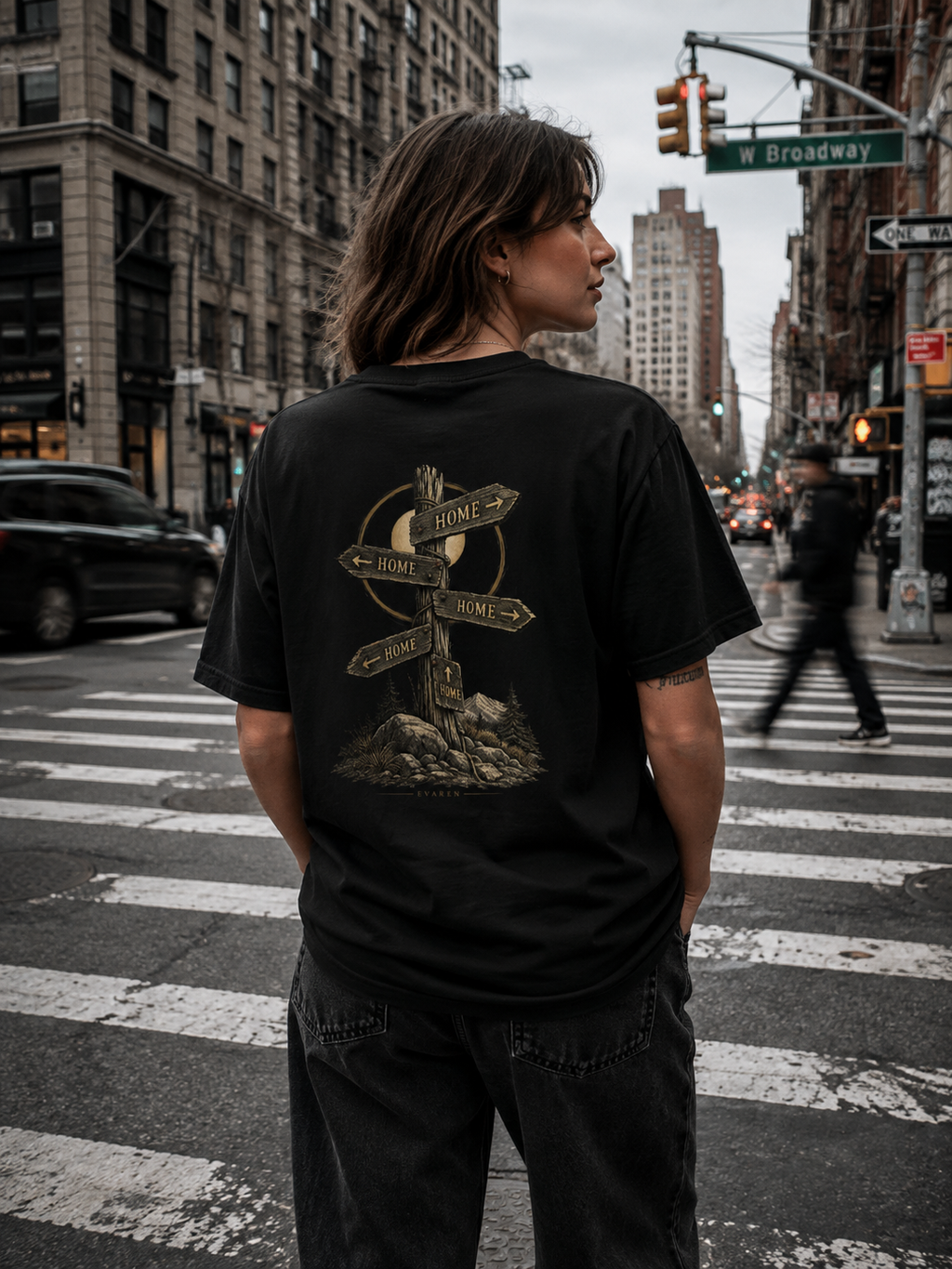 Woman wearing The Way Home Tee by EVAREN in a city intersection, featuring the weathered HOME signpost artwork on the back.