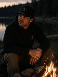 Man wearing a black EVAREN Signature Cap beside a lakeside campfire at dusk