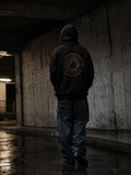 Person wearing The Break Hoodie from behind in a dim concrete urban passage, showing the gold broken-wall back artwork.