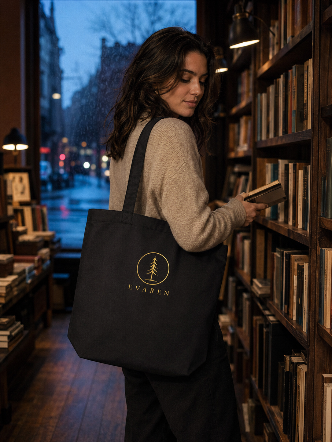 Woman browsing books in a warmly lit bookstore while carrying the black EVAREN organic cotton tote bag with antique-gold evergreen logo.
