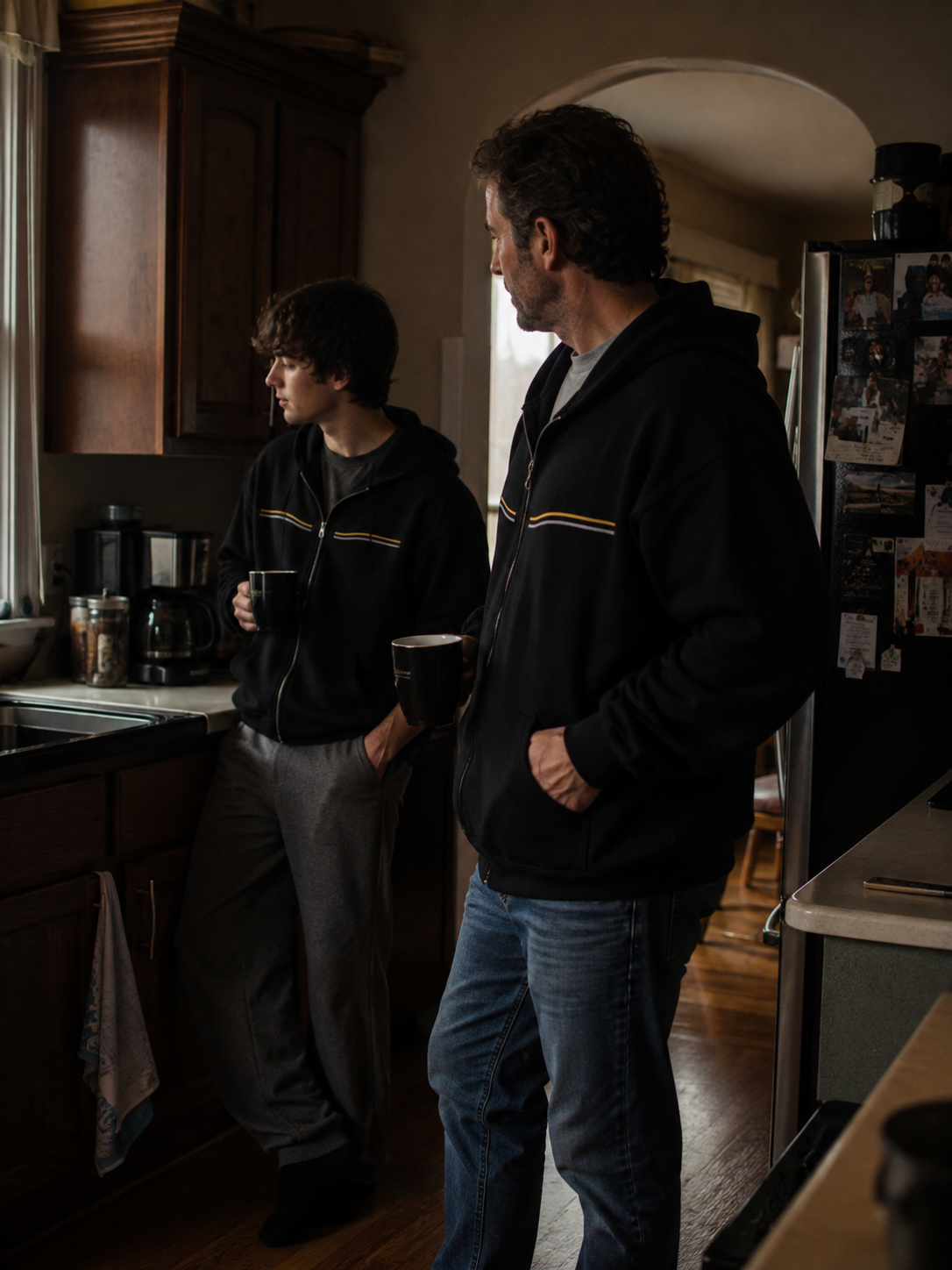 Father and son wearing black EVAREN Side by Side Zip Hoodies together in a lived-in kitchen.