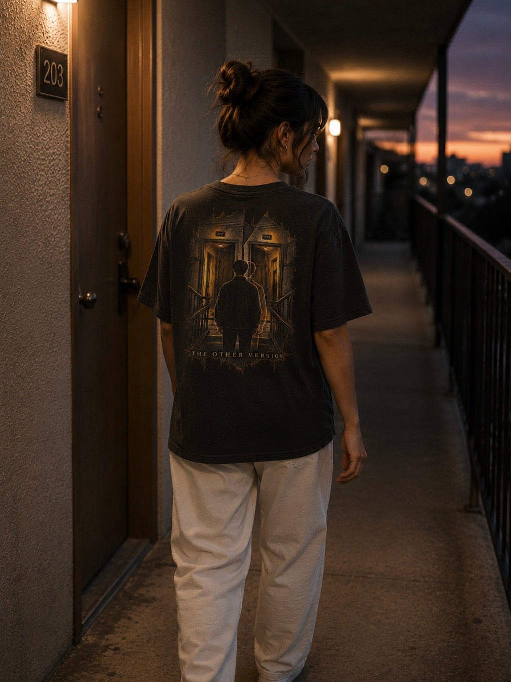 Woman wearing the EVAREN The Other Version Tee while walking through an apartment corridor at dusk, showing the large back artwork.