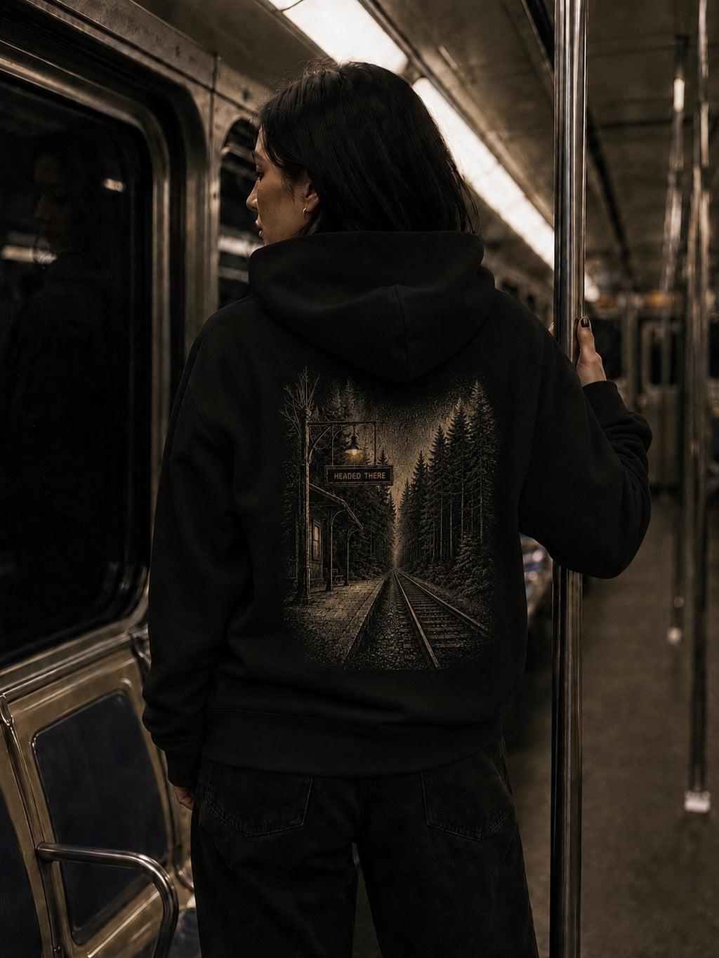 Woman wearing the black EVAREN Headed There Hoodie on a subway, showing the large railway and forest back graphic.