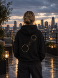 Woman wearing The Necessary Distance Hoodie on a rooftop terrace at dusk, showing the muted gold back artwork.
