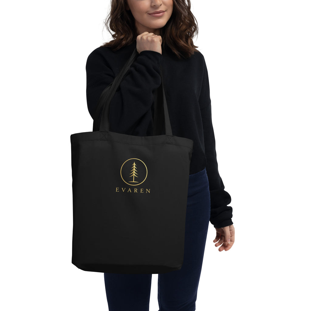 Woman in a black sweatshirt carrying the black EVAREN organic cotton tote bag with antique-gold evergreen logo.