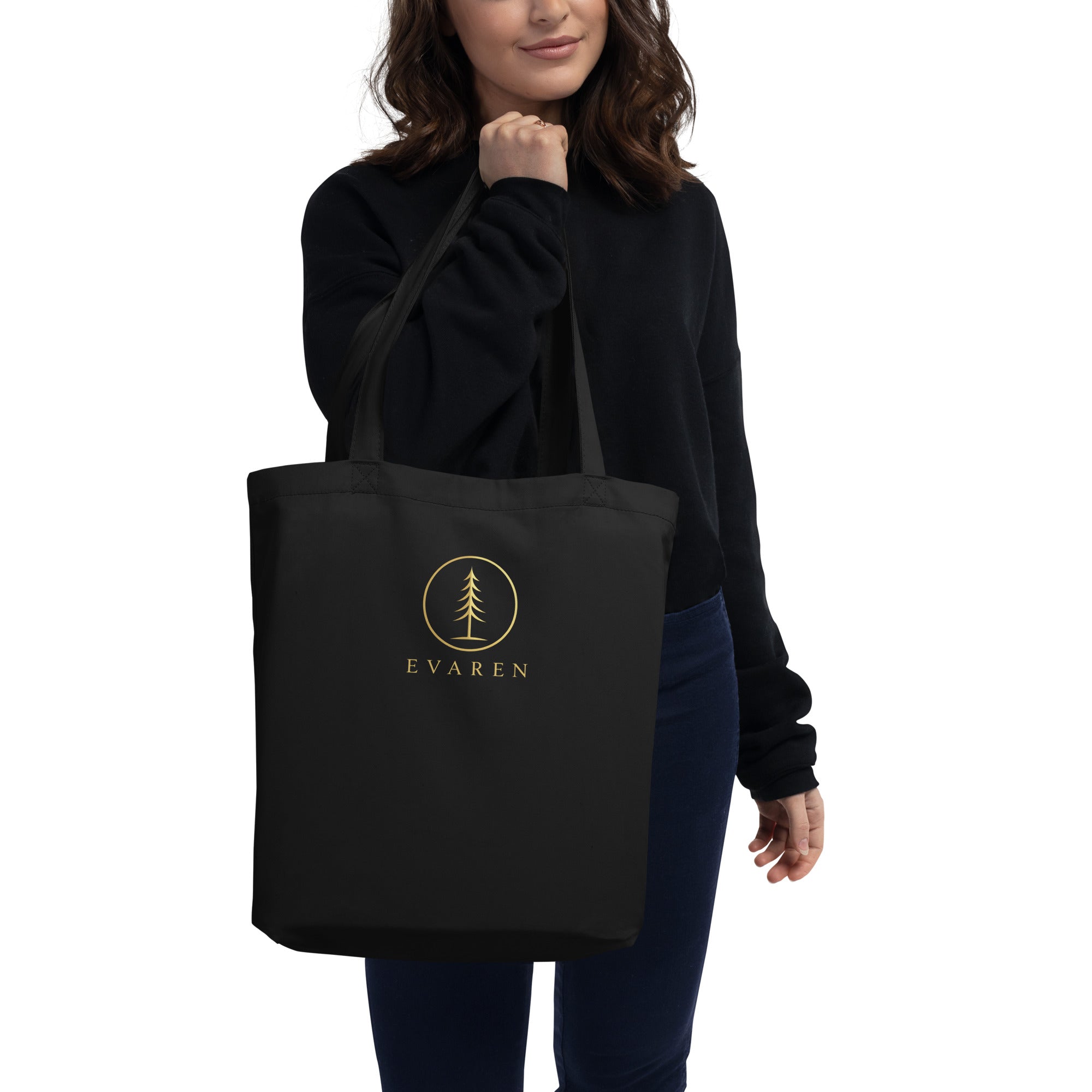 Woman in a black sweatshirt carrying the black EVAREN organic cotton tote bag with antique-gold evergreen logo.