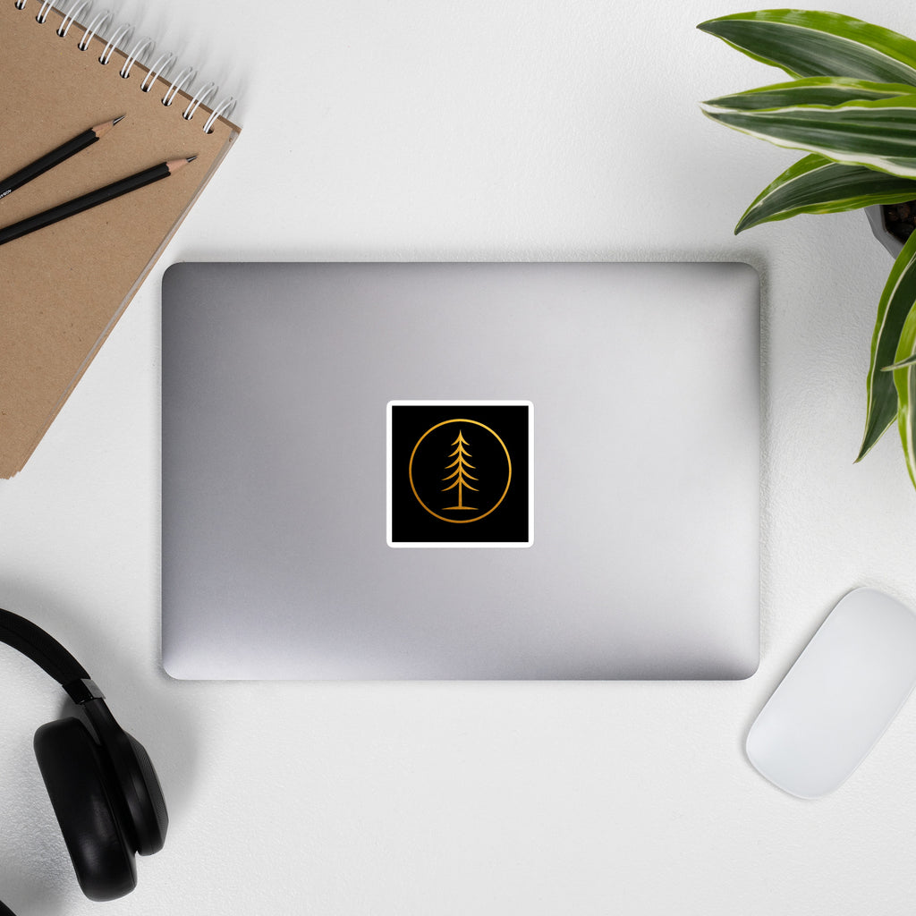 EVAREN square sticker with gold evergreen emblem on a silver laptop at an everyday workspace with a notebook, headphones, mouse, and plant.